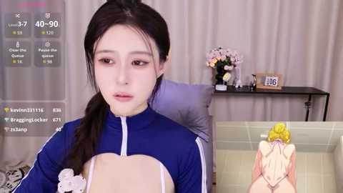 Video of a young East Asian woman with fair skin, long black hair in a ponytail, wearing a blue track jacket. Background includes a beige curtain, a table with flowers, and a digital clock.
