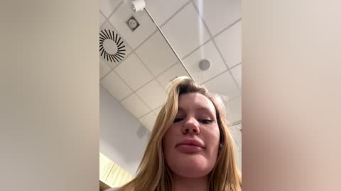 A low-angle video of a smiling, fair-skinned woman with long blonde hair, standing in a bright, modern room with a white ceiling and recessed lights.