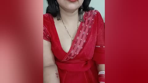 Video of a South Asian woman with medium skin tone wearing a red sari with intricate embroidery, black earrings, and a silver necklace. She has a slight smile and is partially obscured by red curtains.