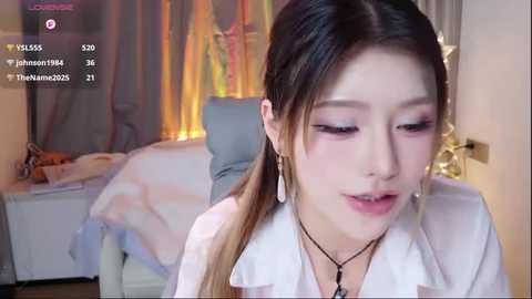 Video of an Asian woman with light skin and long, dark hair in a white blouse, sitting in a dimly lit bedroom with a bed, lamp, and curtains.