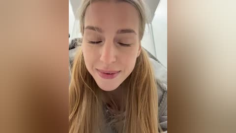 A close-up video of a young woman with fair skin, blonde hair, and closed eyes, wearing a fur-trimmed coat, smiling gently. The background is blurred and neutral-colored, focusing attention on her serene expression.