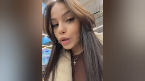 Video of a young woman with long, straight, dark hair, wearing a brown top and a white fur coat. She has a light tan complexion, large eyes, and a small nose piercing. Background shows a grocery store aisle with blue and white packaging.
