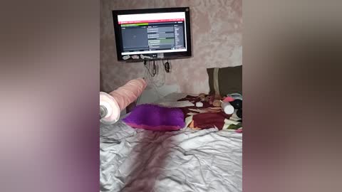 A video of a messy bedroom with a large flat-screen TV mounted on a wall showing a news program. A pink vibrator is charging on the bed, surrounded by disheveled bedding, pillows, and scattered clothes. The background wall has a subtle floral pattern.