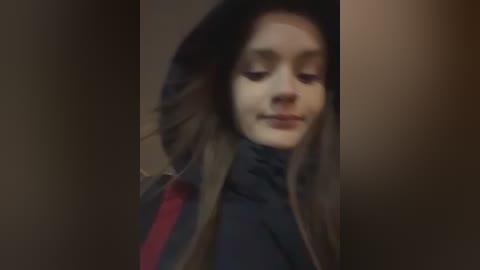 A dimly lit video of a young girl with fair skin, long brown hair, and a dark hooded jacket, looking down, possibly in a corridor.
