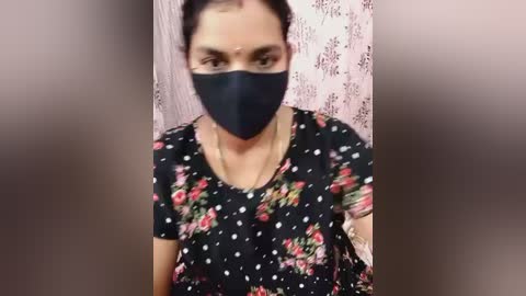 Video of a woman with medium skin tone and dark hair in a ponytail, wearing a black floral dress and black face mask, against a blurred, pink floral-patterned background.