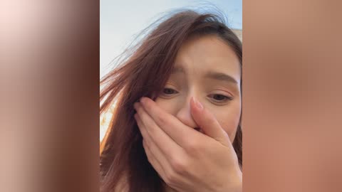 A close-up video of a young Asian woman with long, straight brown hair, covering her mouth with her hand, her expression appearing distressed. The background is blurred with soft lighting.