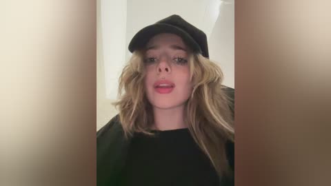 Video of a young Caucasian woman with long, wavy blonde hair, wearing a black beanie and black top, pouting slightly, set against a neutral background.