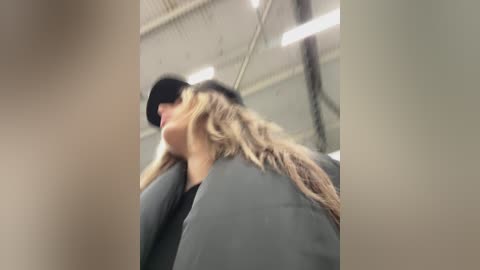 A blurry video of a woman in a black jacket and hat, with long blonde hair, walking in an indoor, industrial setting with high ceilings and fluorescent lights.