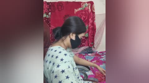 A video of a woman in a white dress with blue stars, wearing a black mask, sitting on a bed covered with a red, floral-patterned sheet.