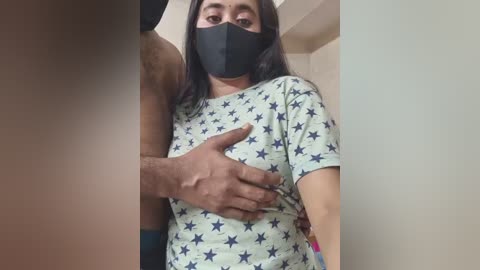 Video of a young woman with medium brown skin, wearing a black face mask, a white t-shirt with blue stars, and a man's hand on her chest. The background is plain beige.