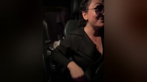 Video of a woman with medium brown skin, wearing glasses, black blazer, and floral blouse, sitting in a dark car, smiling.