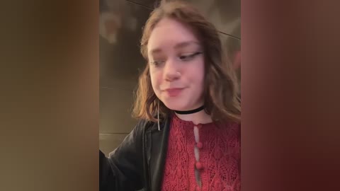 A video of a young woman with light skin, wavy brown hair, and a black choker, wearing a red lace top and black jacket, looking down. The background is blurry, suggesting an indoor setting.