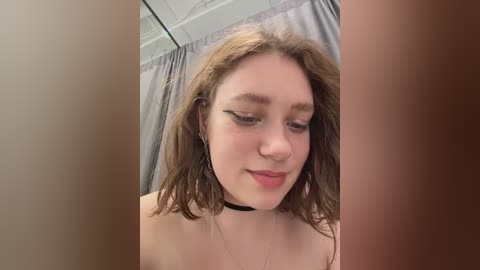 A video of a young woman with shoulder-length wavy brown hair and fair skin, wearing a black choker, and standing in a bathroom with a grey shower curtain.