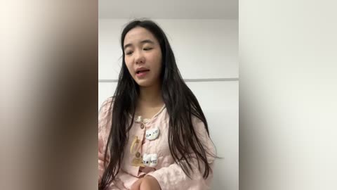 Video of an Asian woman with long black hair, wearing a pink cardigan with white cat-shaped buttons, standing in a white room, slightly out of focus.