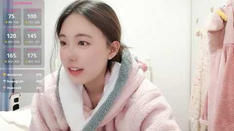 A video of an Asian woman with straight, dark hair, wearing a fluffy pink bathrobe, standing in a modern, minimalist bathroom with white walls and a towel rack.