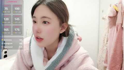 A video of an East Asian woman with straight brown hair, wearing a light pink bathrobe, in a bathroom with a white towel hanging on the door.