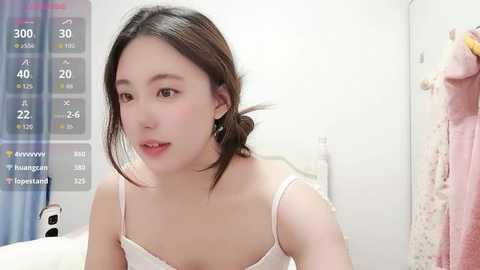 Media: Video of an Asian woman with fair skin, wearing a white spaghetti-strap top, looking thoughtful in a well-lit bedroom with a digital clock displaying 3000, 30, and 40 minutes.