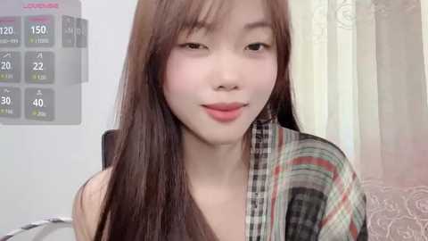 Video of a young Asian woman with long, straight brown hair, fair skin, wearing a plaid jacket, smiling softly, in a light-colored room with a digital thermometer and curtains.