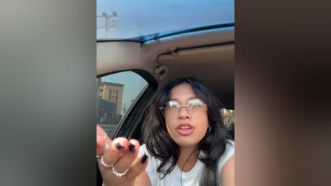 Video of a young Asian woman with long black hair and glasses, wearing a white top, gesturing with her hands while seated in a car, viewed from the backseat.