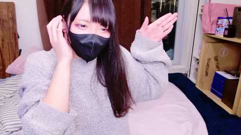 Video of an Asian woman with long black hair, wearing a gray sweater and black face mask, lying on a bed, gesturing with her hands. Background includes a wooden door, a bookshelf, and a pink bag.