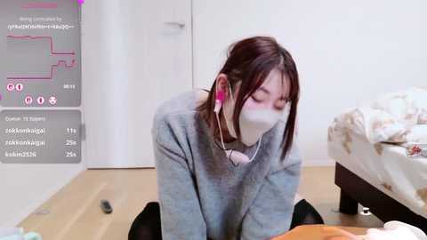 Video of an Asian woman with long dark hair, wearing a gray sweater, white mask, and red headphones, kneeling on a wooden floor in a simple bedroom.