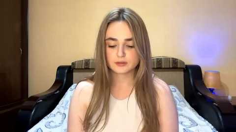 Video of a young woman with long, straight, light brown hair, wearing a sleeveless white top, sitting on a bed with patterned sheets. The room has beige walls and wooden furniture.