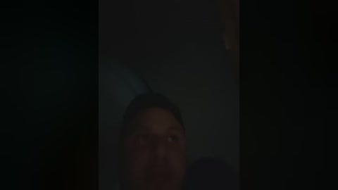 A dimly lit video of a woman with light skin and dark hair, partially visible due to the low lighting and shadows. Her expression is indistinct, with the background blurred and dark, creating an ominous atmosphere.