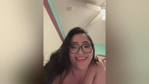 Video of a smiling, topless woman with long dark hair, wearing black-framed glasses, in a room with light walls, a ceiling fan, and a wooden bed visible in the background.
