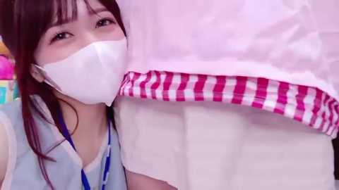 Video of an Asian woman in a hospital gown with a white mask, blue lanyard, and a pink-and-white checked blanket, standing next to a bed.