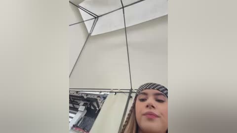 A video of a young woman with light skin, long brown hair, and wearing a black and white checkered headband, standing in a small, narrow, white-walled room with a large mirror.