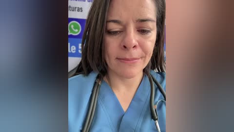A video of a middle-aged woman with light skin and straight brown hair, wearing blue scrubs and a stethoscope, looking down with a concerned expression in a healthcare setting.
