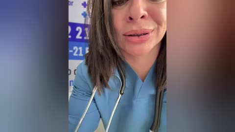 Video of a smiling, light-skinned woman with shoulder-length brown hair, wearing a light blue scrub top. She holds a stethoscope around her neck. Background shows a blurred blue and white sign with numbers.