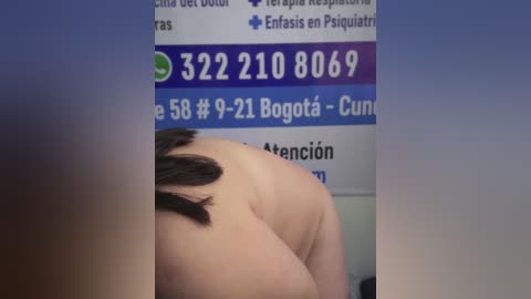 Video of a person with short, dark hair bending over, revealing their back and shoulder area. A blue and white sign with phone numbers and text in Spanish is partially visible in the background.