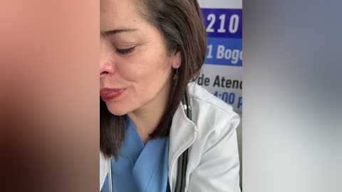 A video showing a female doctor with light skin and straight brown hair, wearing a white lab coat over a light blue shirt, examining a patient's genital area.