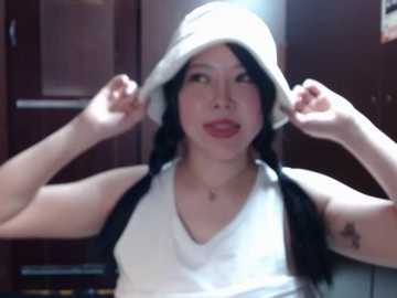 A video of a young Asian woman with light skin, wearing a white bucket hat, black twin braids, and a white tank top, smiling and adjusting her hat, indoors with wooden doors and cabinets in the background.