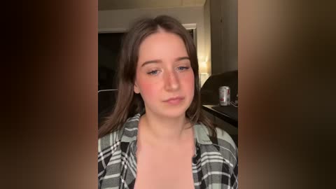 Video of a young woman with fair skin and long brown hair, wearing a plaid shirt, sitting indoors with blurred background.