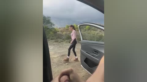 A video of a young woman in a pink top and black leggings walking near an open car door in a dusty, outdoor setting with greenery and a cloudy sky.