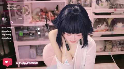 Video of a slender, pale-skinned woman with short black hair and a white top, leaning forward with a slight smile. Background features a cluttered shelf filled with figurines and objects.
