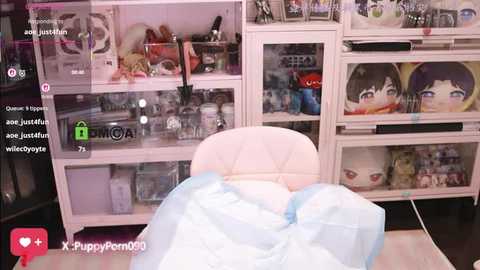 Video of a cozy, pastel-toned room with a plush white chair draped in light blue fabric, featuring a soft toy and a white-framed TV. The background includes a glass-fronted cabinet filled with more plush toys and decorative items.