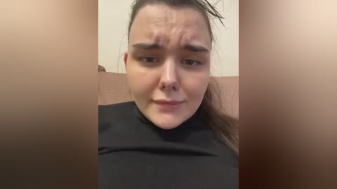 Video of a young woman with fair skin, dark hair tied back, wearing a black turtleneck. She has a confused expression, frowning with furrowed brows, set against a beige wall and a brown couch.