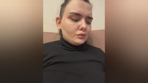 Video of a woman with fair skin, dark hair tied back, wearing a black turtleneck, making a pouty face, set against a beige wall with a pinkish cushion.