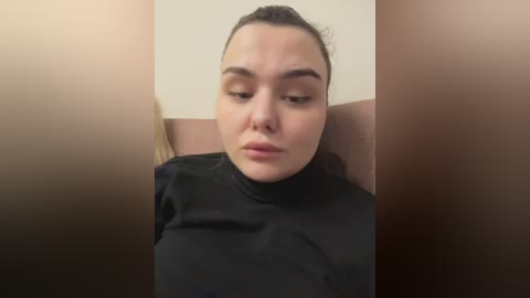 A video shows a young woman with fair skin, dark hair tied back, wearing a black turtleneck, sitting in a beige chair against a beige wall. She looks contemplative.