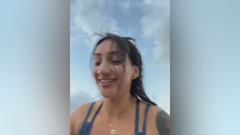Video of a smiling woman with wet, dark hair, wearing a blue bikini top, standing in front of a cloudy sky, slightly out of focus.