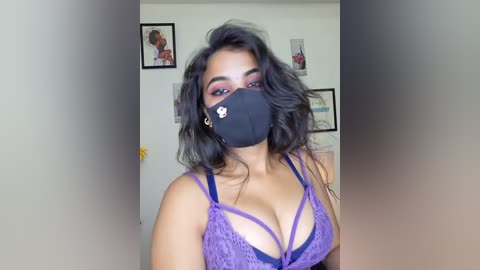 Media: Video of a woman with medium skin tone, wearing a purple lace bra and black face mask, showcasing her medium-sized breasts. Background features framed art and a beige wall.