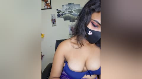 A video of a woman with medium skin tone, wearing a blue bikini top that exposes her ample breasts, black face mask, and black hair. Background includes framed photos, a yellow star, and a black chair.