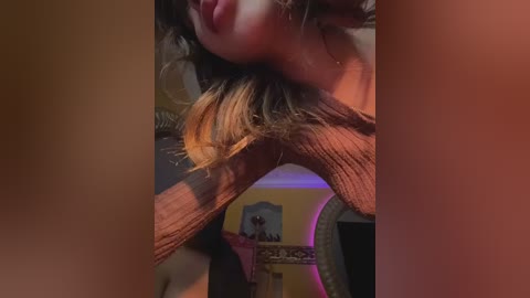 Media: Video of a woman with light brown skin, wearing a brown knit top, leaning over a wooden chair, her hair partially covering her face. Background includes a mirror, framed picture, and a soft purple light.