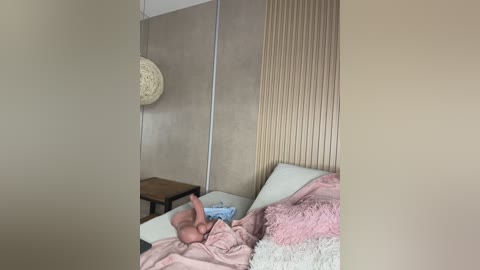 Video of a cozy, modern bedroom with a baby lying on a bed covered in pink blankets, next to a wooden nightstand and textured beige wall.