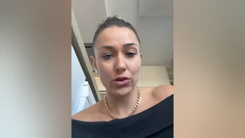 Video of a woman with fair skin, brown hair pulled back, wearing a black off-shoulder top and gold chain necklace. She has a neutral expression and is indoors with beige walls and a white ceiling.