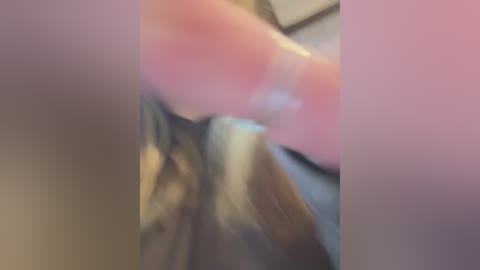 A blurred video featuring a close-up of a hand holding a pink object, possibly a toy, with visible fingers. The background is indistinct, likely a room with neutral-colored walls.