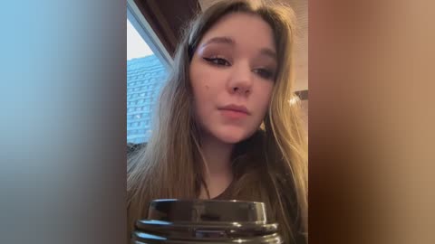 Video of a young Caucasian woman with long, straight blonde hair, wearing black eyeliner, sipping from a glass. Background shows a window with a blue sky and a modern building.